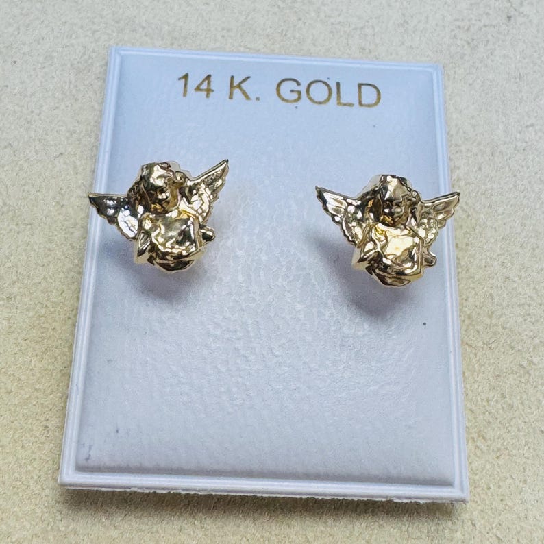 Op de afbeelding: Een paar goudkleurige engel-oorstekers. De oorbellen worden gepresenteerd op een witte kaart met de tekst "14 K. GOLD" in gouden letters. De oorbellen hebben gedetailleerde engelenfiguren met vleugels.