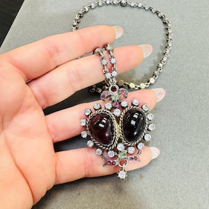 Może przedstawiać: Srebrny naszyjnik z sercowatym wisiorem, na którym znajdują się dwa ciemnofioletowe kamienie otoczone przez przezroczyste i różowe cyrkonie. Wisiorek zdobi filigranowy wzór w srebrze i mała kokardka.