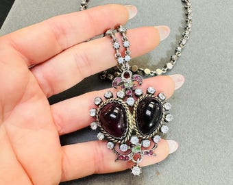 May include: A silver necklace with a heart-shaped pendant featuring two dark purple stones surrounded by clear and pink rhinestones. The pendant is detailed with a silver filigree design and a small bow.
