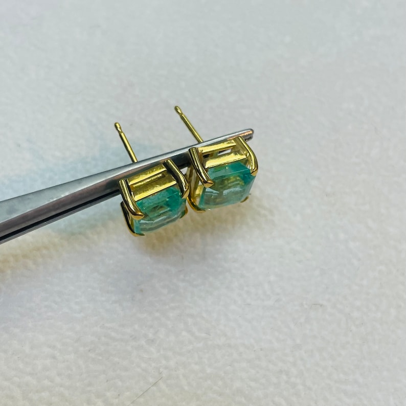 May include: A pair of gold earrings with emerald green gemstones. The earrings are square shaped and have a simple design.