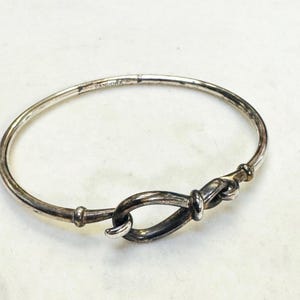 May include: A silver-toned Pandora bracelet with a knot design. The bracelet is circular with a knot-shaped clasp. The metal has a slightly aged appearance. The bracelet is likely made of sterling silver or a similar metal.
