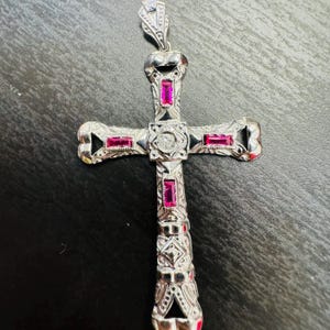 May include: A silver cross pendant with intricate detailing and pink gemstones. The cross is adorned with a diamond-shaped center stone.