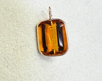 Op de afbeelding: Een rechthoekige hanger met een grote, gefacetteerde, amberkleurige edelsteen. De edelsteen is gezet in een goudkleurig metalen frame met een kleine lus aan de bovenkant voor een ketting. De hanger heeft een warme uitstraling.