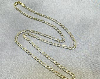 May include: A gold-toned Figaro chain necklace featuring a repeating pattern of three small circular links followed by one elongated oval link. The metal chain has a polished finish and a lobster claw clasp at the end, displaying a classic link design.