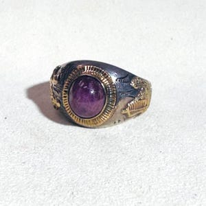 May include: A silver ring with a large, oval, purple gemstone set in a gold bezel. The ring has gold accents on the sides with detailed designs. The ring is on a white surface.