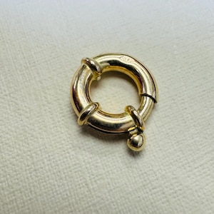 May include: A circular gold spring ring clasp featuring a polished metallic finish. The jewelry component has a small protruding knob used to operate the spring-loaded mechanism, allowing the ring to open and close securely for attaching chains or pendants.