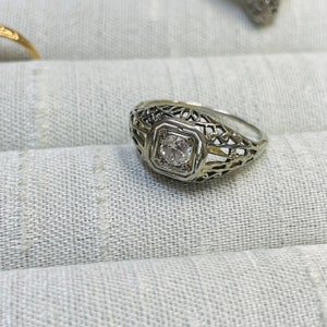 May include: A silver filigree ring with a single round diamond in the center.