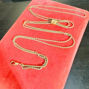Könnte beinhalten: Eine goldfarbene Kette mit einem dekorativen Verschluss und einem kleinen, quadratischen Anhänger mit einem sternförmigen Design. Die Kette liegt auf einem roten Samtstoff.
