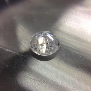 May include: A clear, round, faceted gemstone with a slight gray tint. The gemstone is sitting on a silver surface.