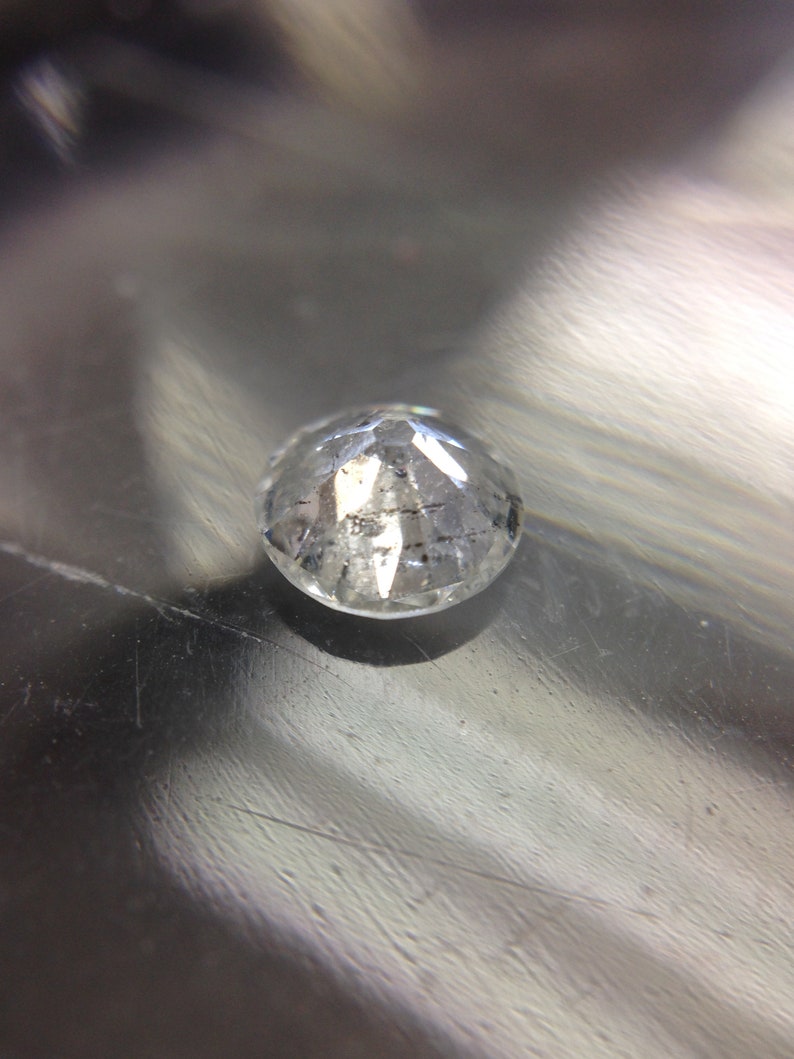 May include: A clear, round, faceted gemstone with a slight gray tint. The gemstone is sitting on a silver surface.