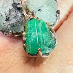 Puede incluir: Un anillo con un cristal de esmeralda en bruto de color verde vibrante en un engaste de plata junto a una piedra preciosa en bruto más grande de color verde menta pálido. El engaste metálico tiene un aspecto texturizado y orgánico.