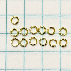 May include: A close-up of 14 gold jump rings. The rings are arranged in two rows on a white gridded surface.
