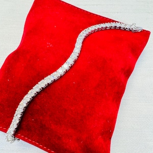 May include: A silver bracelet with numerous round, clear gemstones. The bracelet is displayed on a red velvet surface, showcasing its sparkle and design. The clasp is visible at the end of the bracelet.