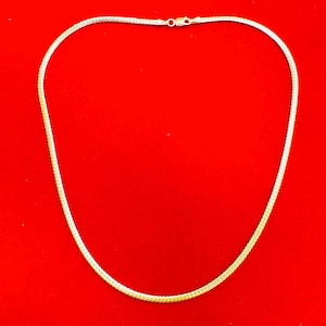 May include: A gold-coloured chain necklace with a woven design. The necklace is displayed against a bright red background. The chain has a small clasp at the top.
