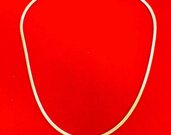 Pode incluir: Um colar de corrente dourada com um design tecido. O colar é exibido em um fundo vermelho brilhante. A corrente tem um pequeno fecho na parte superior.