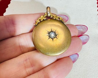 May include: Antique gold-tone pendant with a central starburst design and a small, clear gemstone. The pendant has an ornate, decorative bail. The round pendant is held in a hand.