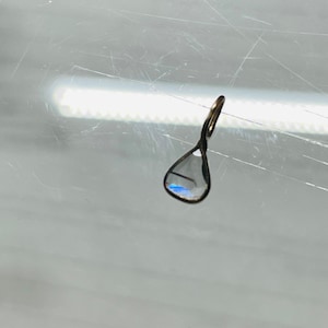 May include: A teardrop-shaped pendant with a clear glass stone that has a blue iridescent sheen. The pendant is made of silver and has a loop at the top for attaching to a chain.