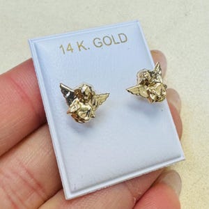 Op de afbeelding: Een paar goudkleurige engel-oorstekers. De oorbellen worden gepresenteerd op een witte kaart met de tekst "14 K. GOLD" in gouden letters. De oorbellen hebben gedetailleerde cherubijnontwerpen met vleugels.