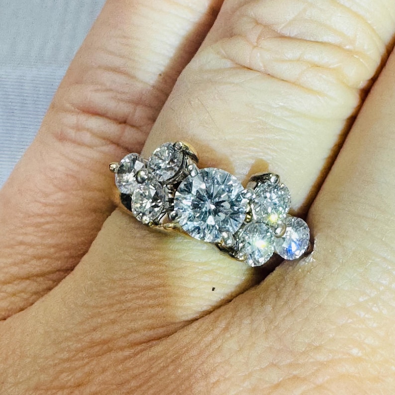 May include: A cluster ring featuring a large, round, clear diamond as the focal point, surrounded by smaller, sparkling diamonds. The ring has a gold band and is set on a finger, showcasing its brilliance.