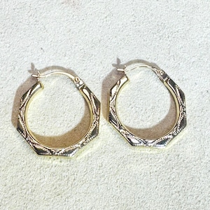 Op de afbeelding: Een paar goudkleurige oorringen. Elke oorring heeft een achthoekige vorm met een gestructureerd, patroonontwerp langs de randen. De oorbellen worden gepresenteerd op een lichtgekleurd oppervlak.