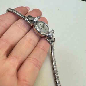 K&ouml;nnte beinhalten: Eine silberne Uhr mit einem runden Zifferblatt und einem silbernen Armband. Das Armband besteht aus einer Reihe kleiner, miteinander verbundener Kreise. Die Uhr hat ein wei&szlig;es Zifferblatt mit schwarzen r&ouml;mischen Ziffern als Stundenmarkierungen und schwarzen Zeigern. Auf jeder Seite des Zifferblatts befinden sich zwei kleine Diamanten.