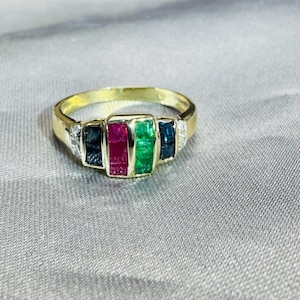 May include: A gold ring featuring a central row of four rectangular channel-set gemstones in dark blue, red, and green, flanked by two small clear stones on each side. The band is polished gold, showcasing a multi-colored gemstone arrangement.