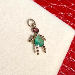 Peut inclure: Un pendentif doré en forme de personne. La tête est une petite pierre précieuse rose et ronde, et le corps est une pierre précieuse verte en forme de goutte d'eau. De petites perles dorées forment les bras et les jambes. Le pendentif est sur une surface blanche.