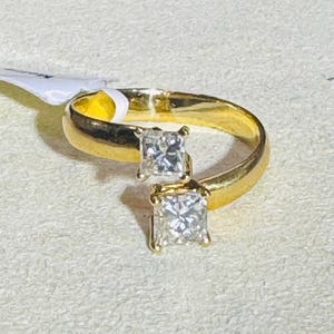 Puede incluir: Un anillo abierto de oro con dos piedras transparentes de corte cuadrado en engaste de garras. La banda es lisa y pulida, con las dos piedras situadas en los extremos abiertos del anillo, creando un diseño cruzado con un acabado brillante.
