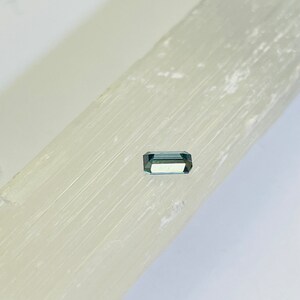 May include: A small, rectangular, green gemstone with a slight blue hue. The gemstone is sitting on a white, textured surface.