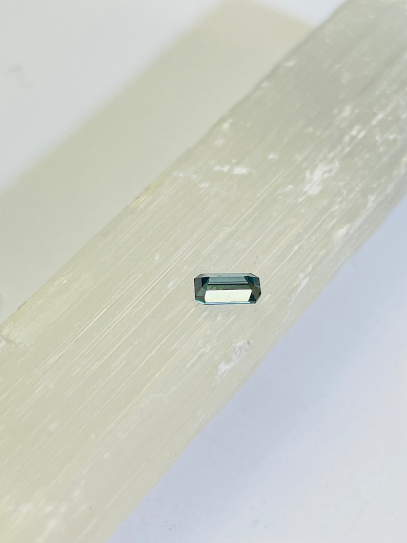 May include: A small, rectangular, green gemstone with a slight blue hue. The gemstone is sitting on a white, textured surface.
