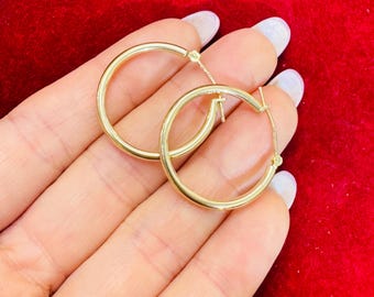 May include: A pair of gold hoop earrings. The earrings are round and have a polished finish. The hoops are held in a hand against a red background.