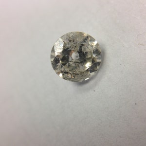 May include: A clear, round, faceted gemstone with a hole in the center. The gemstone has a slightly cloudy appearance.