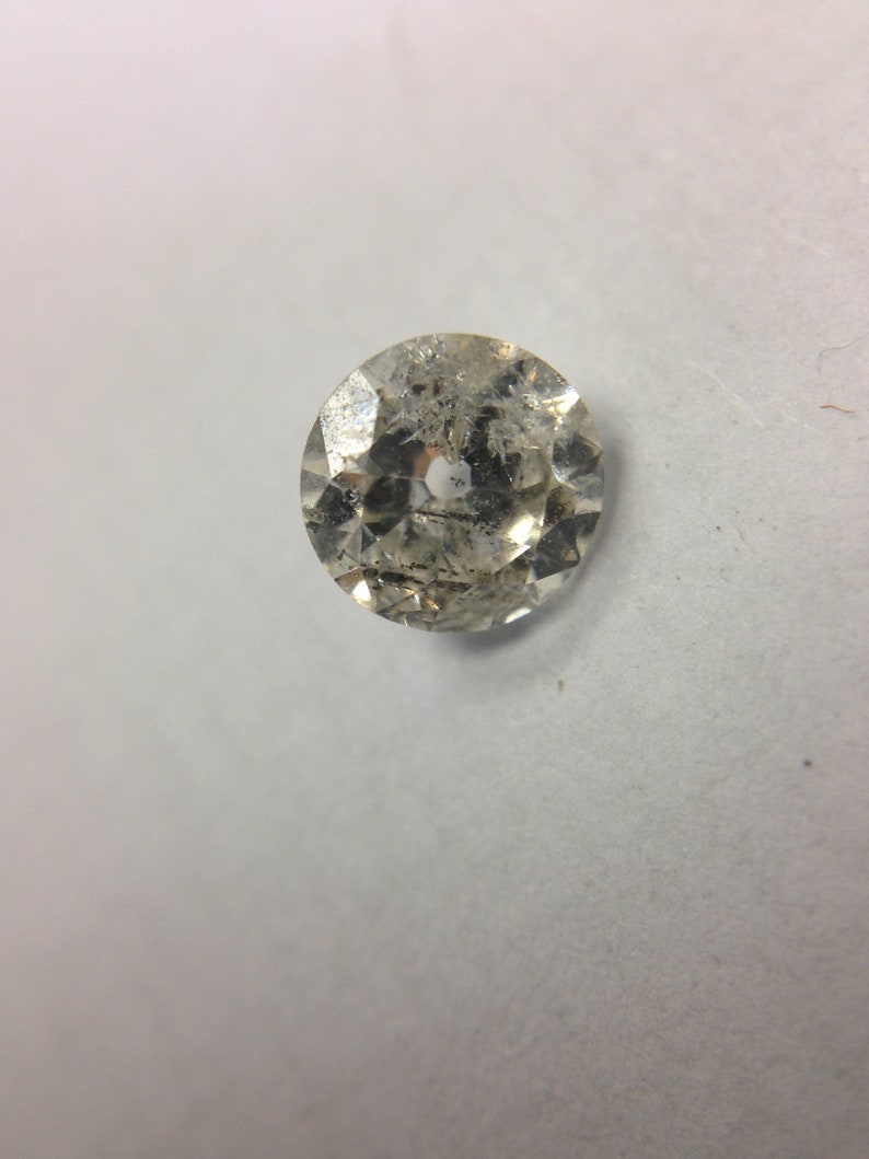 May include: A clear, round, faceted gemstone with a hole in the center. The gemstone has a slightly cloudy appearance.