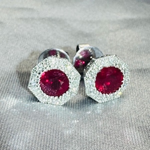 May include: A pair of stud earrings featuring a central round-cut deep red gemstone surrounded by a halo of small, clear, sparkling stones. The setting is octagonal in shape, providing a geometric frame for the vibrant red center stone.