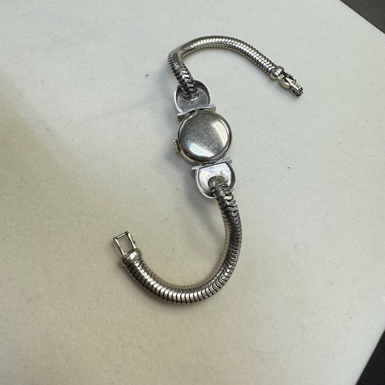 K&ouml;nnte beinhalten: Eine silberne Uhr mit einem runden Zifferblatt und einem Schlangenkettenarmband. Die Uhr hat ein wei&szlig;es Zifferblatt und schwarze Zeiger.