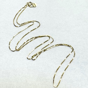 May include: A thin, gold-toned chain necklace featuring a delicate, flat, square-link design. The necklace includes a small, circular spring-ring clasp at one end, showcasing a reflective, metallic finish throughout the entire length of the chain.