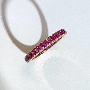 May include: A rose gold ring set with numerous small, vibrant red gemstones. The ring is a full eternity band, with the gemstones encircling the entire band. The ring is displayed on a white surface, with a shadow cast to the side.