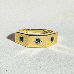May include: A gold-colored ring with a geometric design. The ring features three dark blue gemstones set within star-shaped frames. The ring has a flat, octagonal band. The ring is displayed on a light-colored surface.