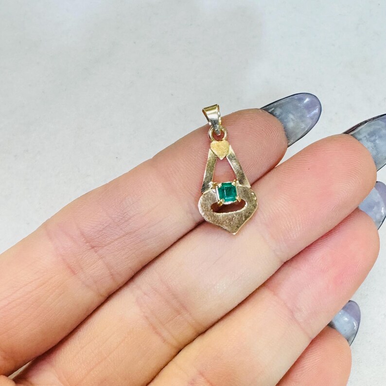 May include: Gold-toned pendant with a heart-shaped top and a teardrop design. The pendant features a square, emerald-green gemstone at its center. The pendant is suspended from a small loop for a chain.