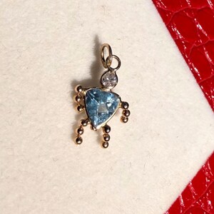 Peut inclure: Pendentif doré orné d'une pierre précieuse bleue en forme de cœur. Au-dessus de la pierre bleue, une petite pierre transparente. De petites perles dorées s'étendent des côtés et du bas du pendentif.