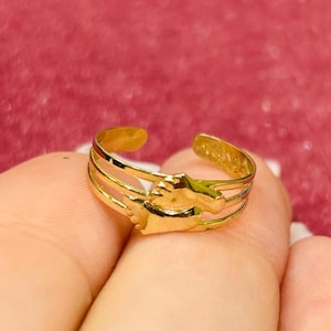 Puede incluir: Un anillo dorado con un diseño de tres bandas. El anillo presenta un diseño calado detallado con un pequeño elemento decorativo. El anillo se muestra en un dedo sobre un fondo rosa. El anillo es probablemente de oro.