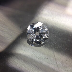 May include: A clear round diamond gemstone with facets, set on a silver surface.