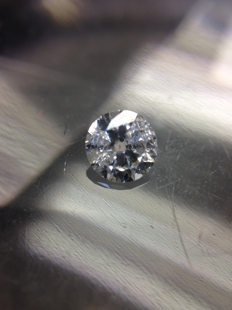 May include: A clear round diamond gemstone with facets, set on a silver surface.