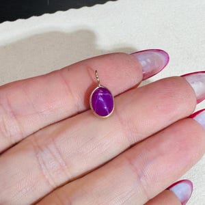 Può includere: Un piccolo pendente ovale con una vibrante pietra preziosa viola. La pietra ha un effetto a stella ed è incastonata in una montatura in metallo dorato. Il pendente è progettato per essere indossato come un ciondolo per collana.