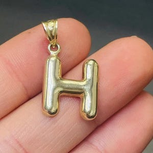 May include: Gold-toned pendant charm in the shape of the letter H. The letter is thick and has a rounded, almost inflated appearance.