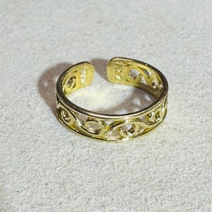 Puede incluir: Un anillo abierto de color dorado que presenta un patrón calado continuo de formas arremolinadas interconectadas. La banda tiene un acabado metálico pulido y una abertura visible en la parte superior, lo que permite ajustar el tamaño.