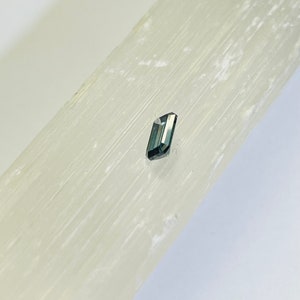 May include: A small, dark gray, faceted gemstone on a white selenite slab.