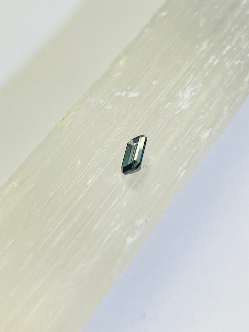 May include: A small, dark gray, faceted gemstone on a white selenite slab.