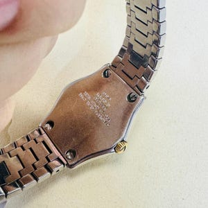 Pode incluir: Um rel&oacute;gio prateado com caixa de a&ccedil;o inoxid&aacute;vel e um detalhe dourado. A pulseira do rel&oacute;gio &eacute; feita de elos de metal interligados. O mostrador do rel&oacute;gio n&atilde;o &eacute; vis&iacute;vel. A parte de tr&aacute;s do rel&oacute;gio tem o texto "STAINLESS STEEL CASE".