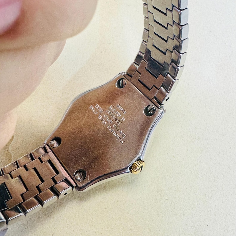 Pode incluir: Um rel&oacute;gio prateado com caixa de a&ccedil;o inoxid&aacute;vel e um detalhe dourado. A pulseira do rel&oacute;gio &eacute; feita de elos de metal interligados. O mostrador do rel&oacute;gio n&atilde;o &eacute; vis&iacute;vel. A parte de tr&aacute;s do rel&oacute;gio tem o texto "STAINLESS STEEL CASE".